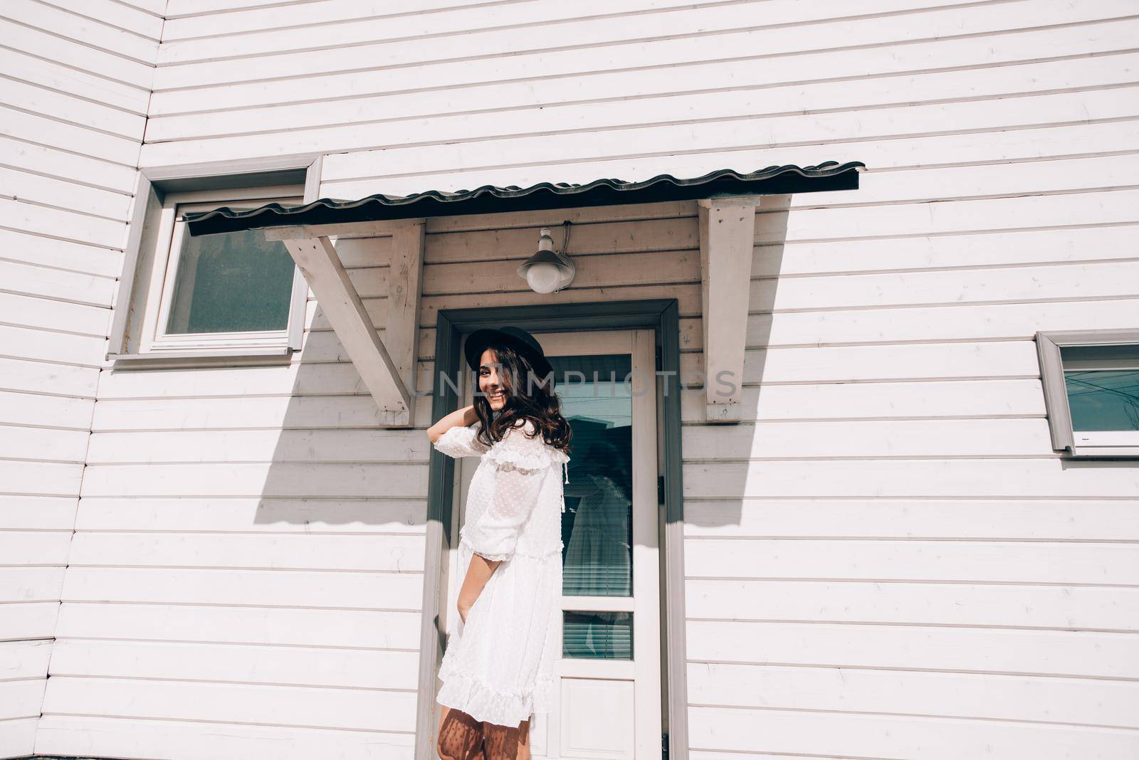 Sunny lifestyle fashion portrait of young stylish hipster woman walking on the street, wearing trendy white dress, black hat and boots. White wooden house on a backgrond.