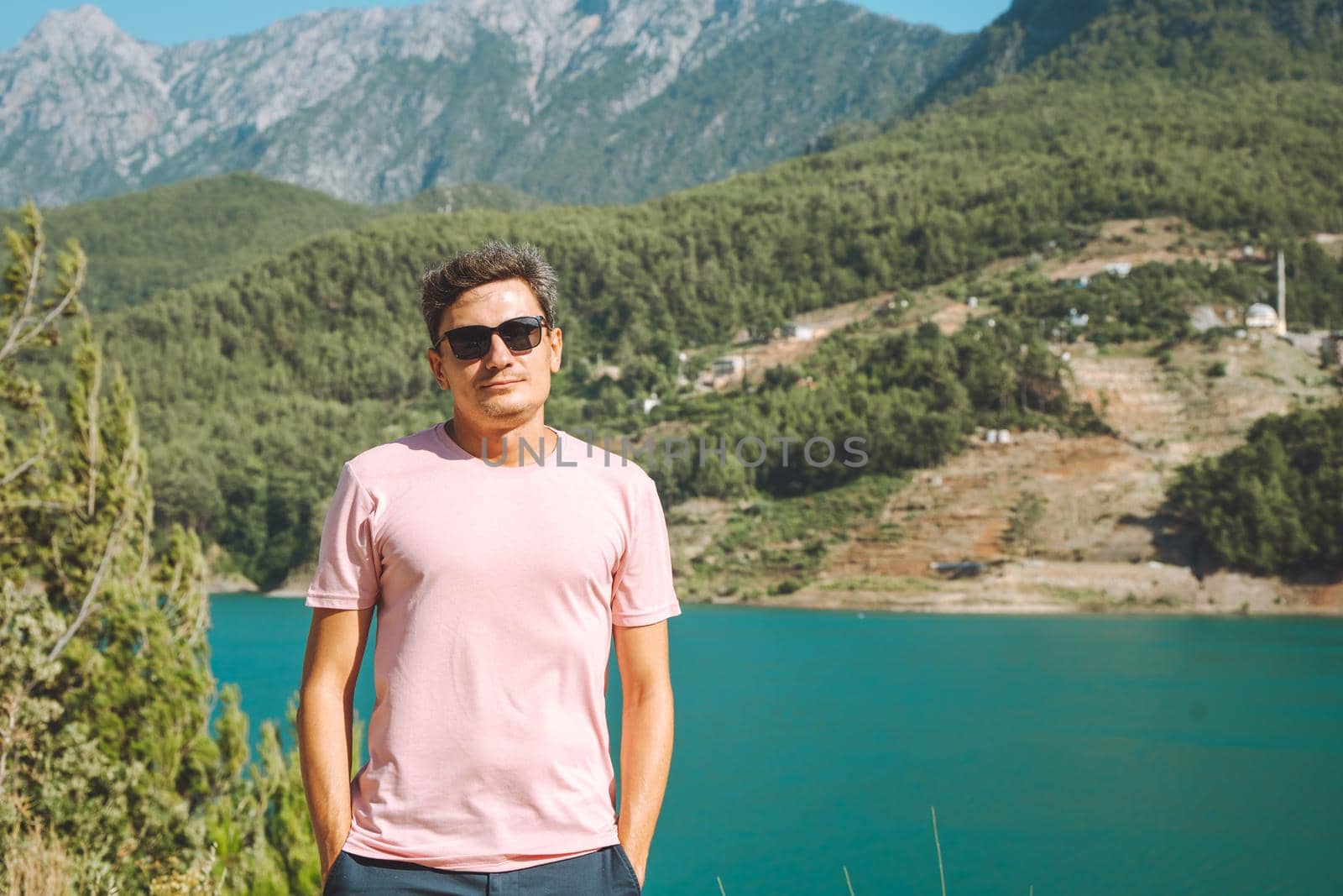 Portrait of Smiling man in sunglasses standing near mountains lake on background. Positive young male traveling on blue lake outdoors travel adventure vacation.