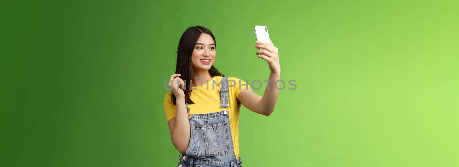 Glamour tender teenage asian female taking selfie, checking haircut, look smartphone front camera, photographing, record video message post online, smiling lovely, green background.