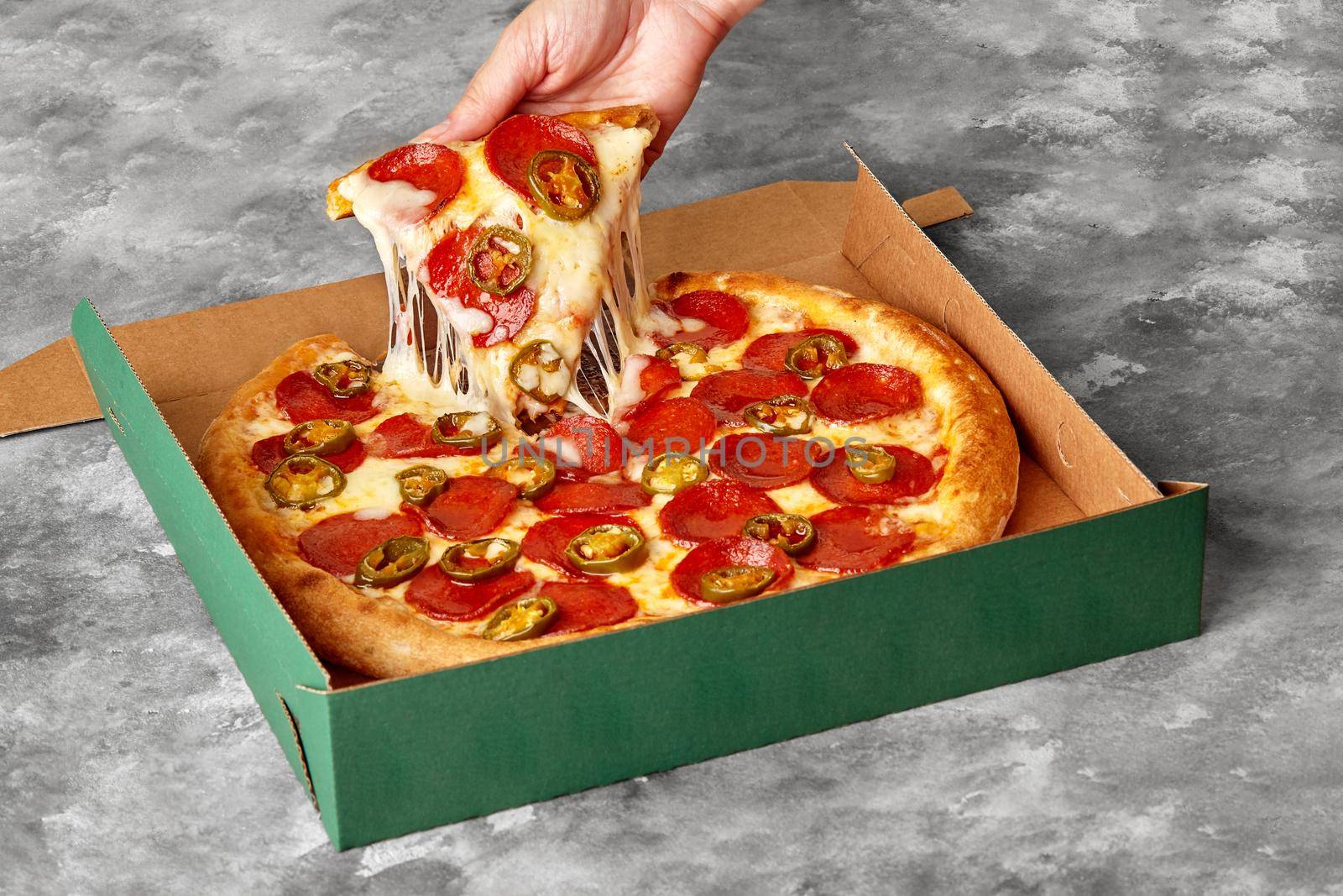 Woman taking slice of thin hot pizza with melted cheese, pepperoni and pieces of spicy jalapeno from cardboard box lying on gray stone table, cropped image. Takeaway food concept