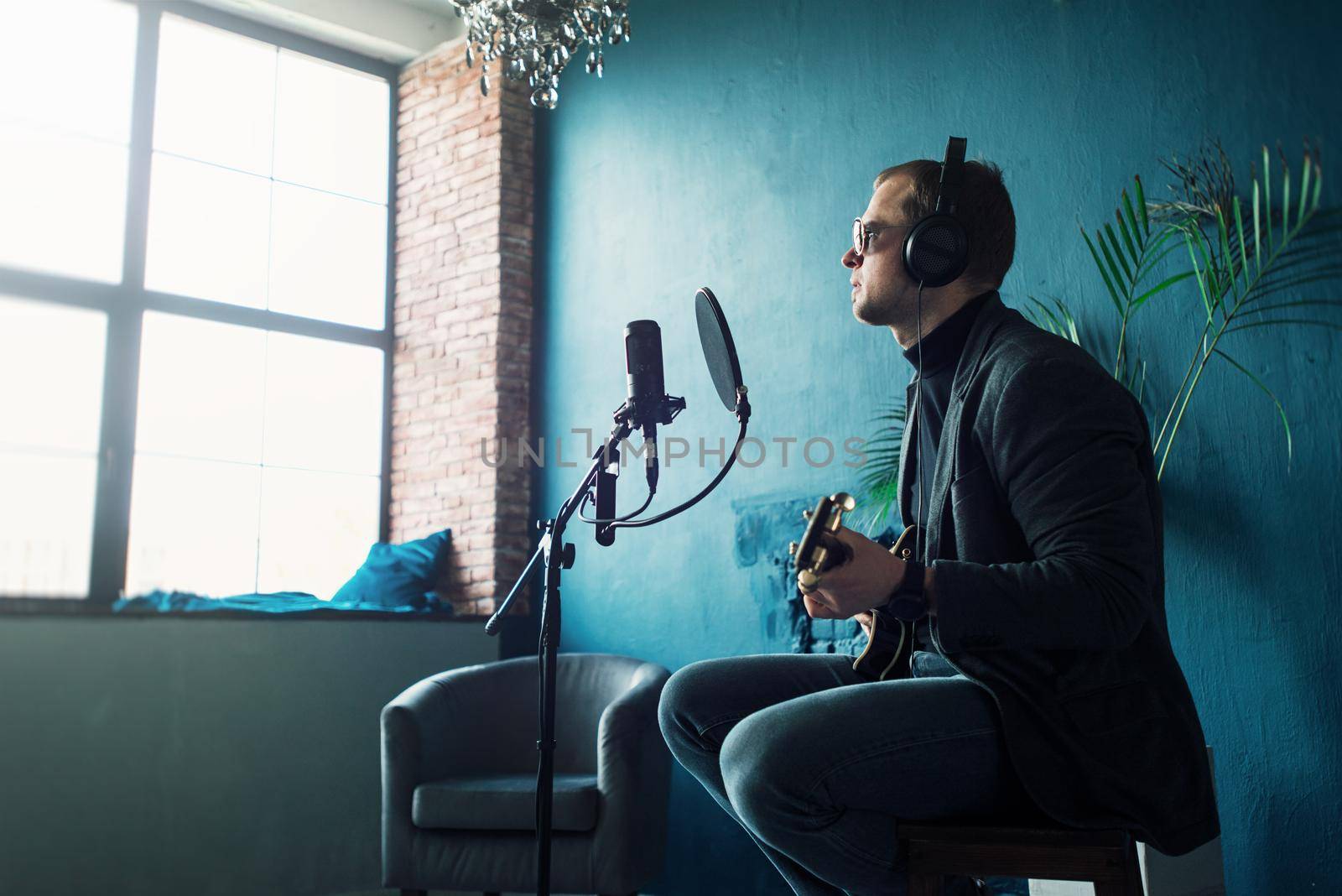 Close up of a man singer sitting on a stool in a headphones with a guitar recording a track in a home studio by Ashtray25