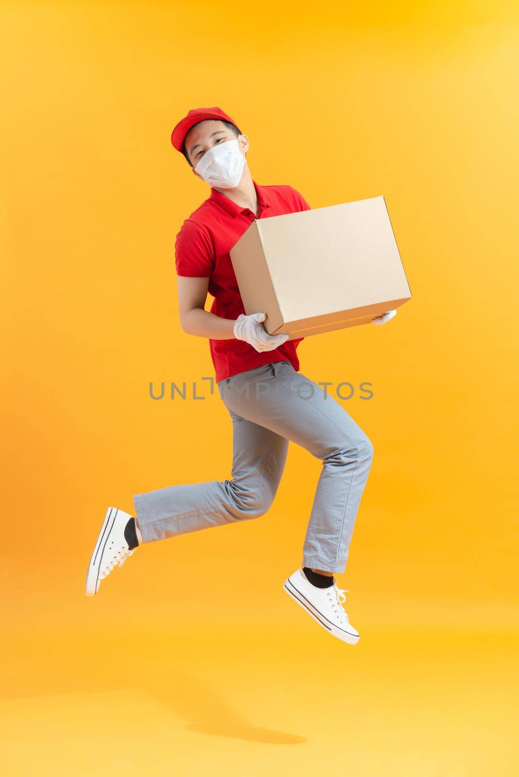 Asian red capped courier wearing face mask and gloves carrying cardboard box and jumping by makidotvn