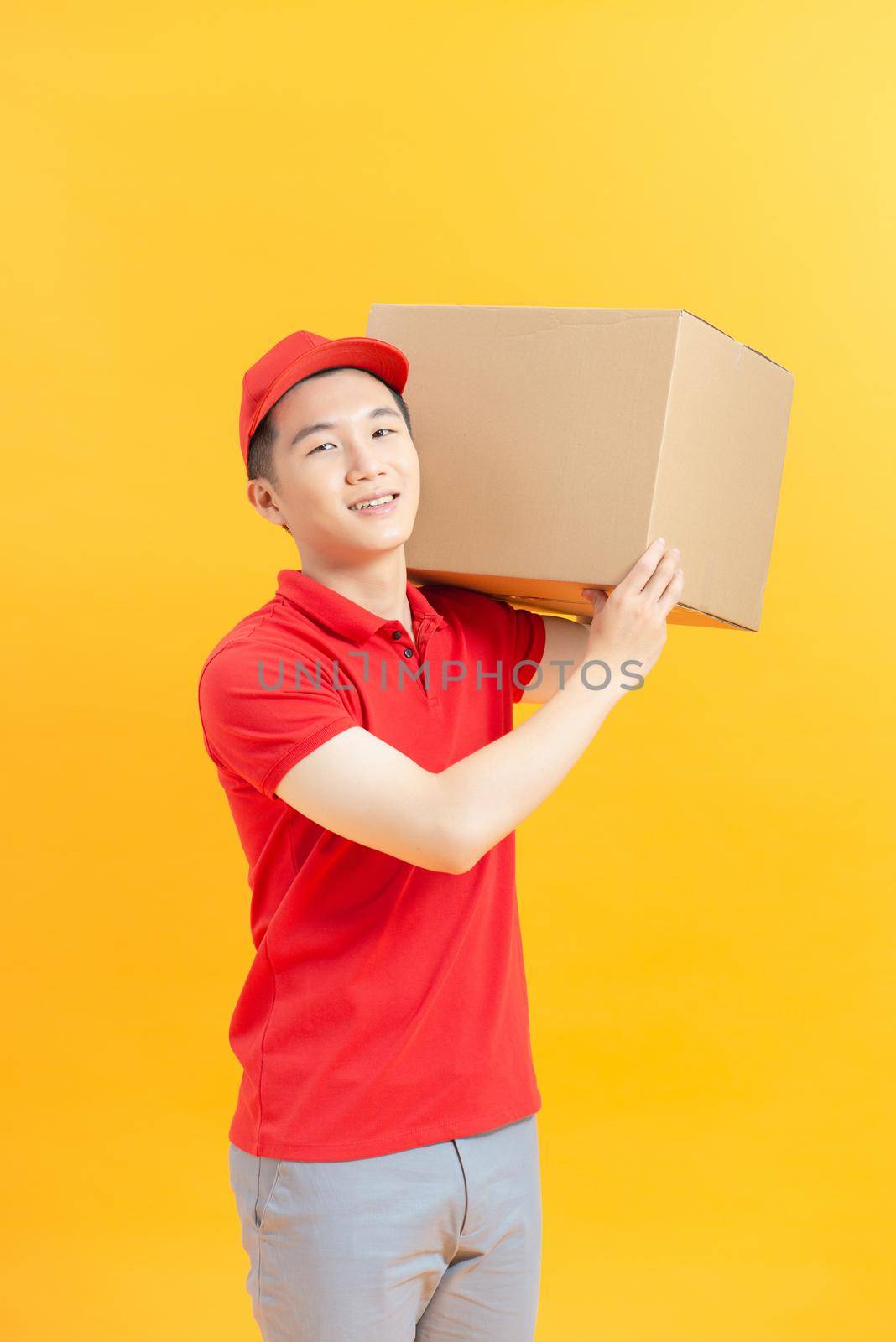 Young smiling logistic delivery man in red uniform holding the box on color background by makidotvn