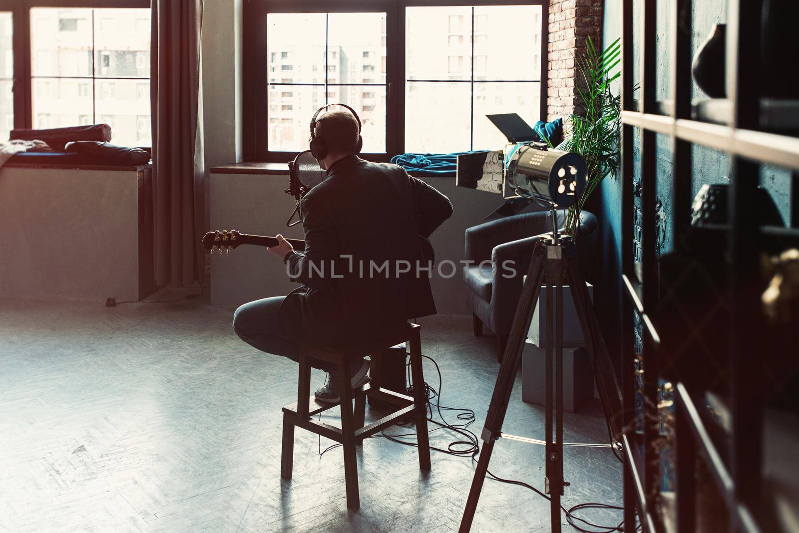 Man singer sitting on a stool in a headphones with a guitar recording a track in a home studio by Ashtray25
