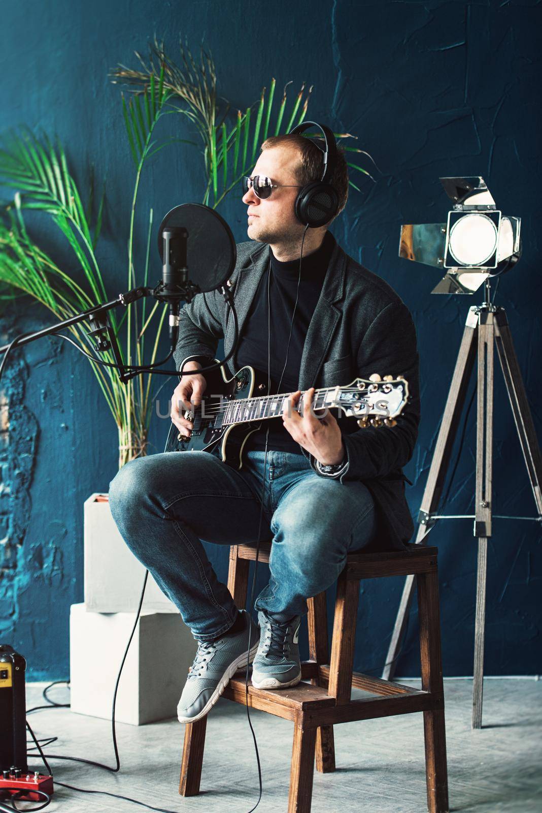 Close up of a man singer in a headphones with a guitar recording a track in a home studio. Man wearing sunglasses, jeans, black shirt and a jacket. side view