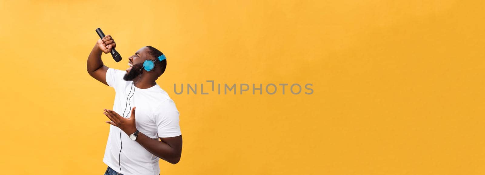 Portrait of cheerful positive chic. handsome african man holding microphone and having headphones on head listening music singing song enjoying weekend vacation isolated on yellow background.