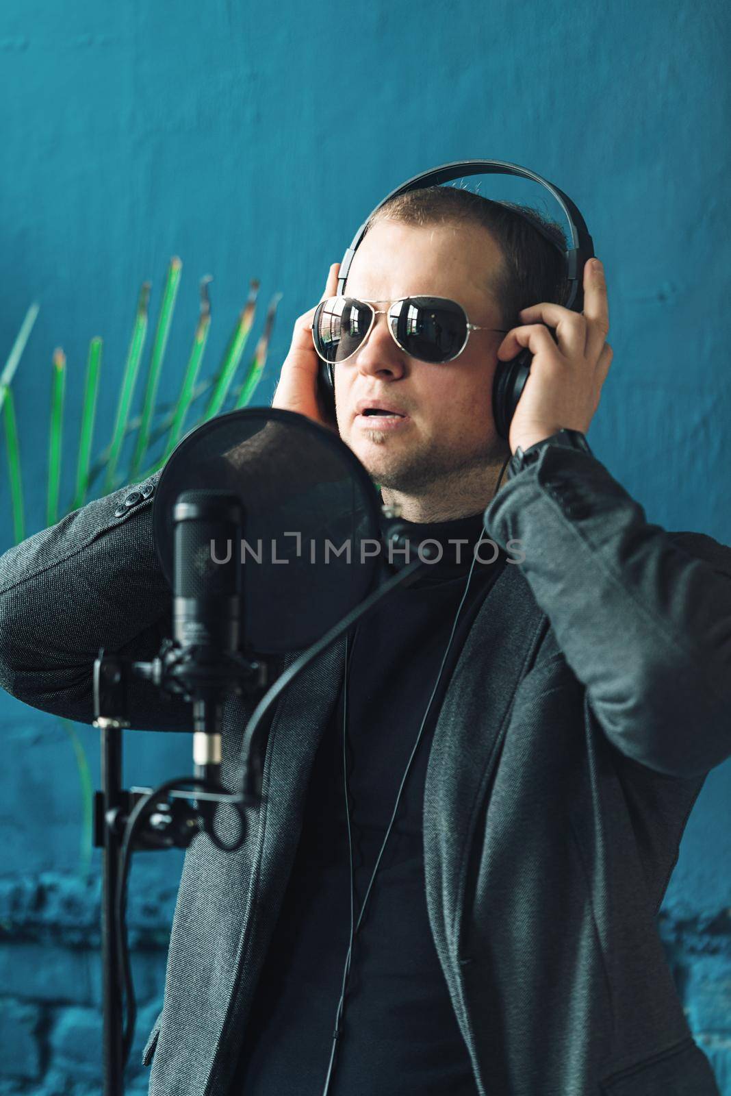 Close up of a man singer in a headphones recording a song in a home studio. Man wearing sunglasses, black shirt and a jacket. side view