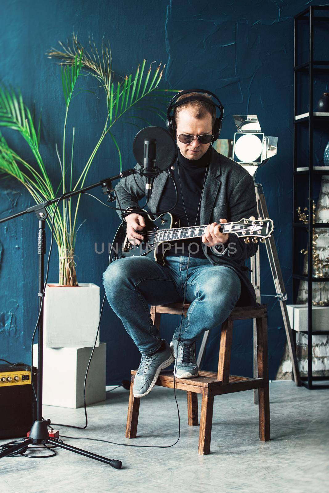 Close up of a man singer in a headphones with a guitar recording a track in a home studio. Man wearing sunglasses, jeans, black shirt and a jacket.