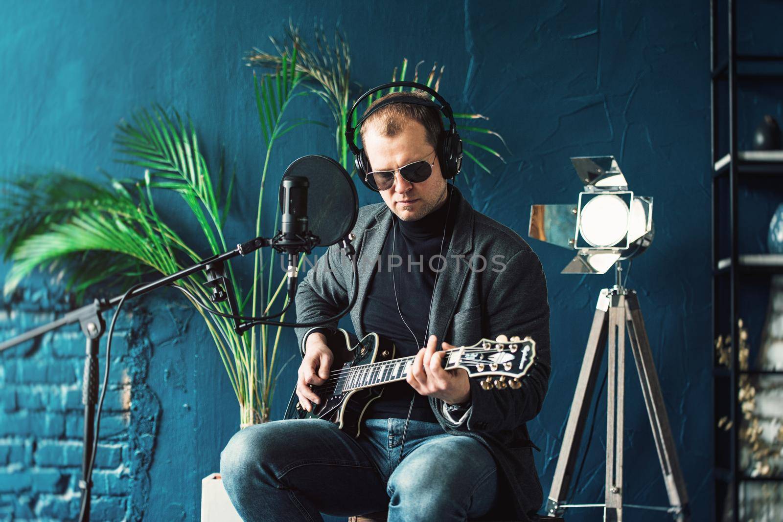 Close up of a man singer in a headphones with a guitar recording a track in a home studio. Man wearing sunglasses, jeans, black shirt and a jacket.
