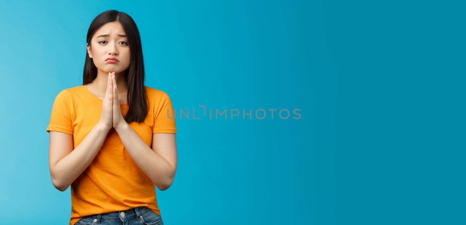 Upset cute silly asian girl praying, plead for help, pouting frowning need, make pitty face, hold hands pray begging for favor, apologizing feelings guilty sad, stand blue background.