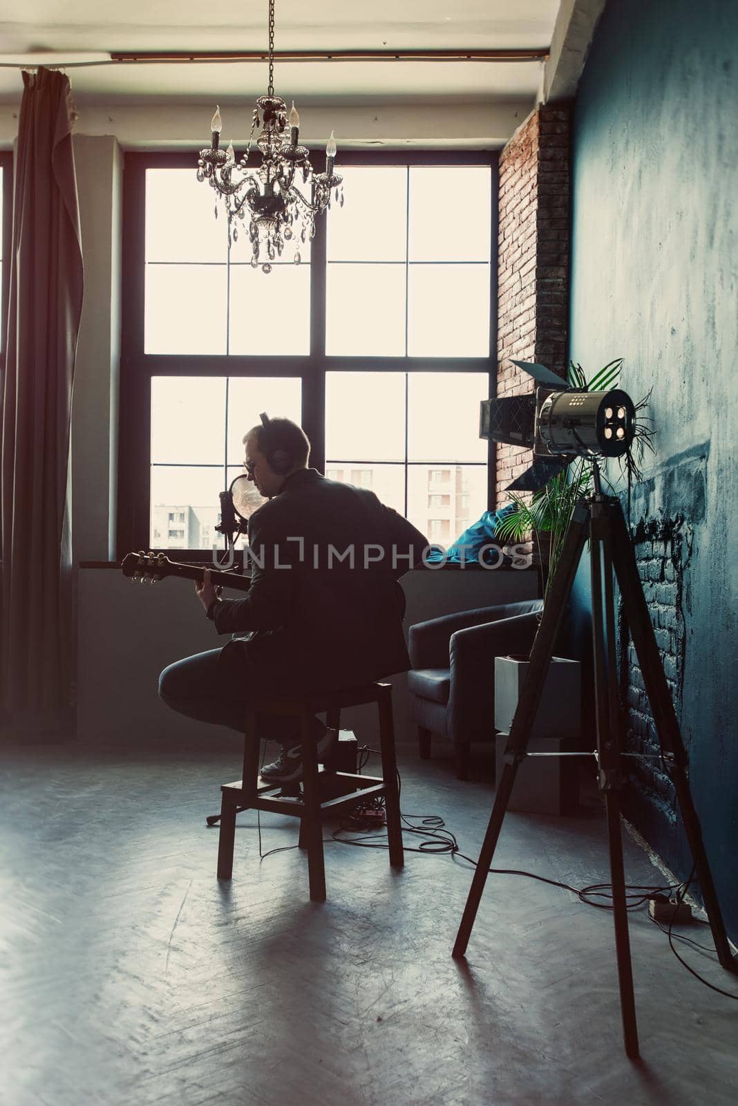 Close up of a man singer in a headphones with a guitar recording a track in a home studio. Man wearing sunglasses, jeans, black shirt and a jacket. Look from back, window on a bakground