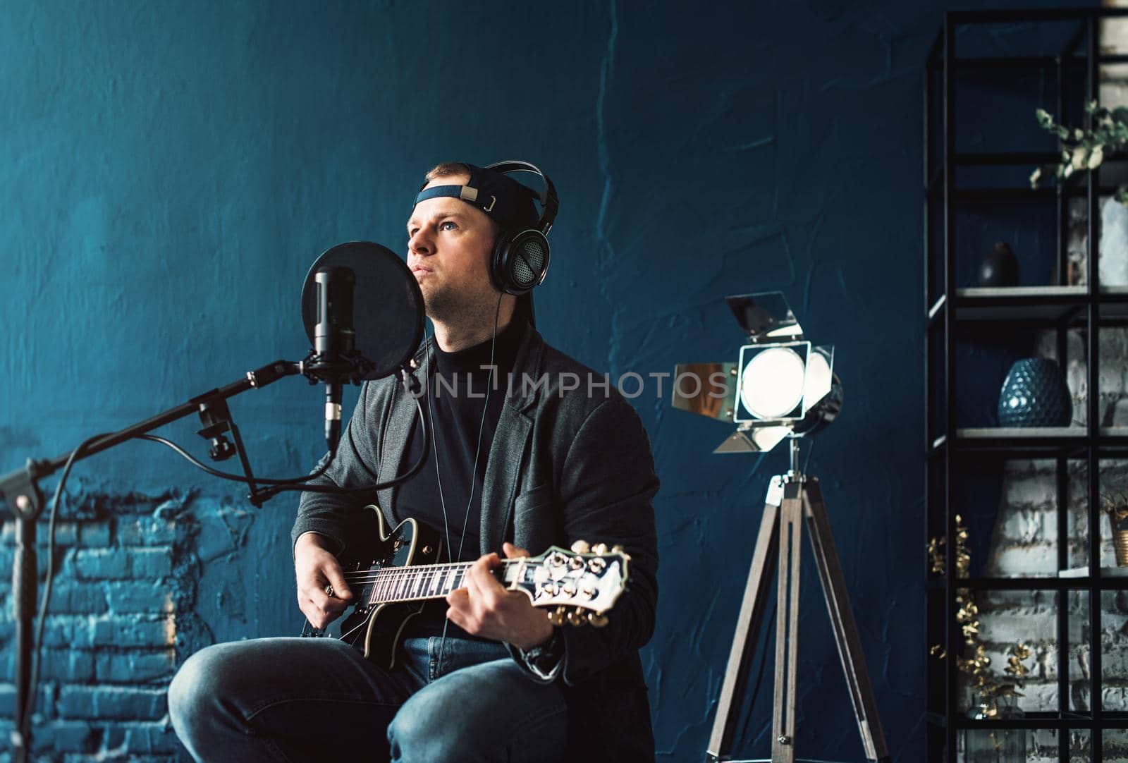 Close up of a man singer sitting on a stool in a headphones with a guitar recording a track in a home studio by Ashtray25