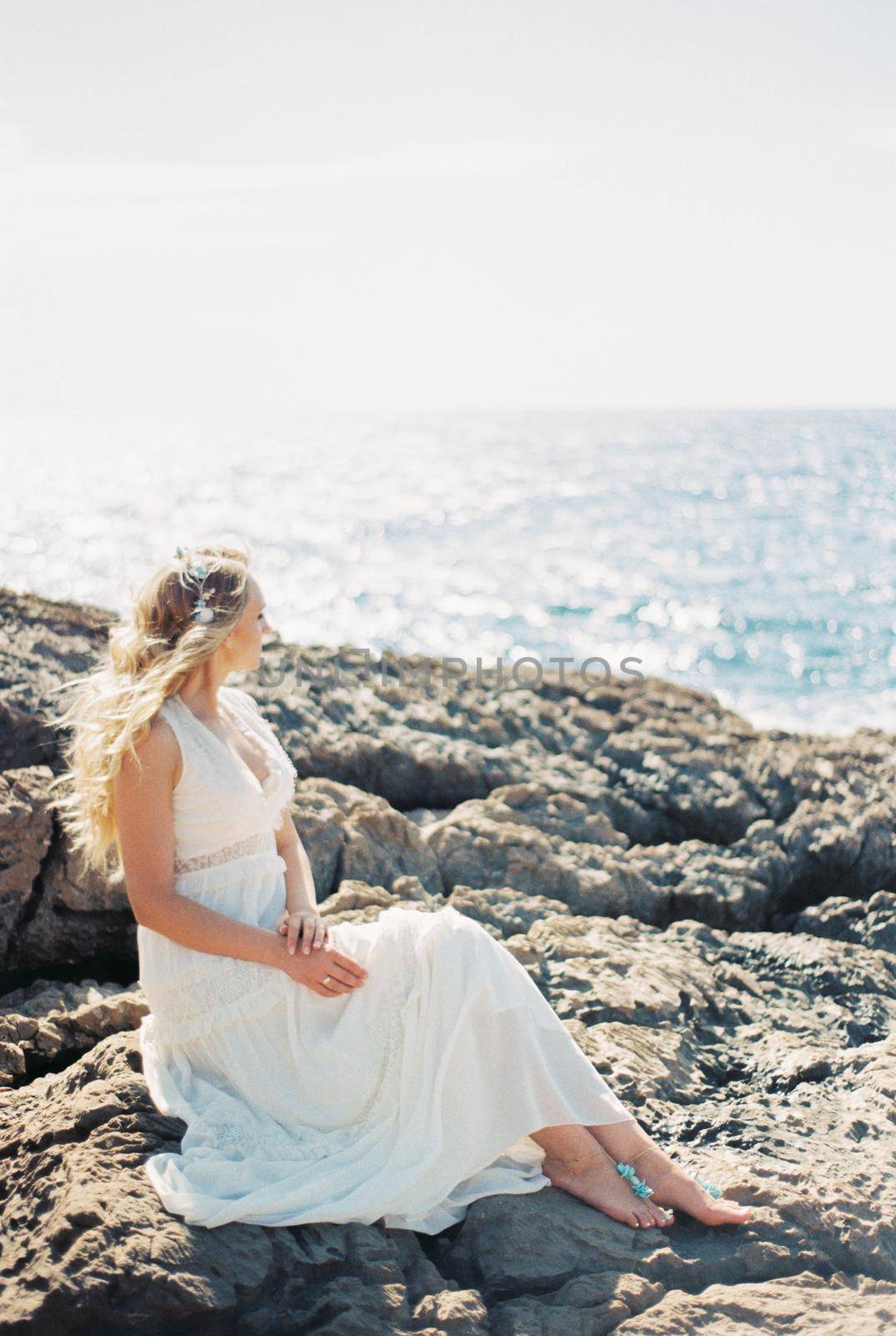 Barefoot bride sits on the rocks and looks at the sparkling sea. High quality photo