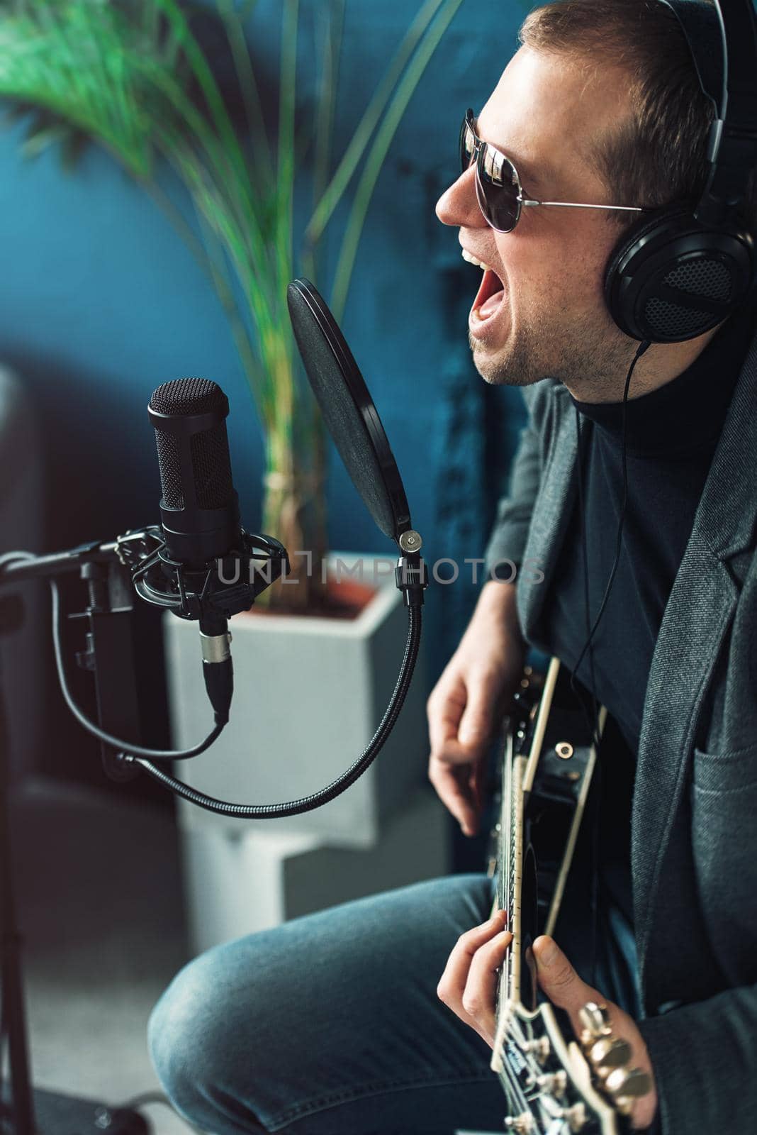 Close up of a man singer in a headphones with a guitar recording a track in a home studio. Man wearing sunglasses, jeans, black shirt and a jacket. side view