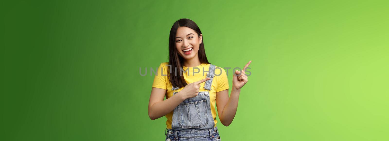 Amused friendly easygoing asian woman having fun discuss funny comedy performance, pointing left smiling laughing carefree, give positive feedback new cafe, stand green background.