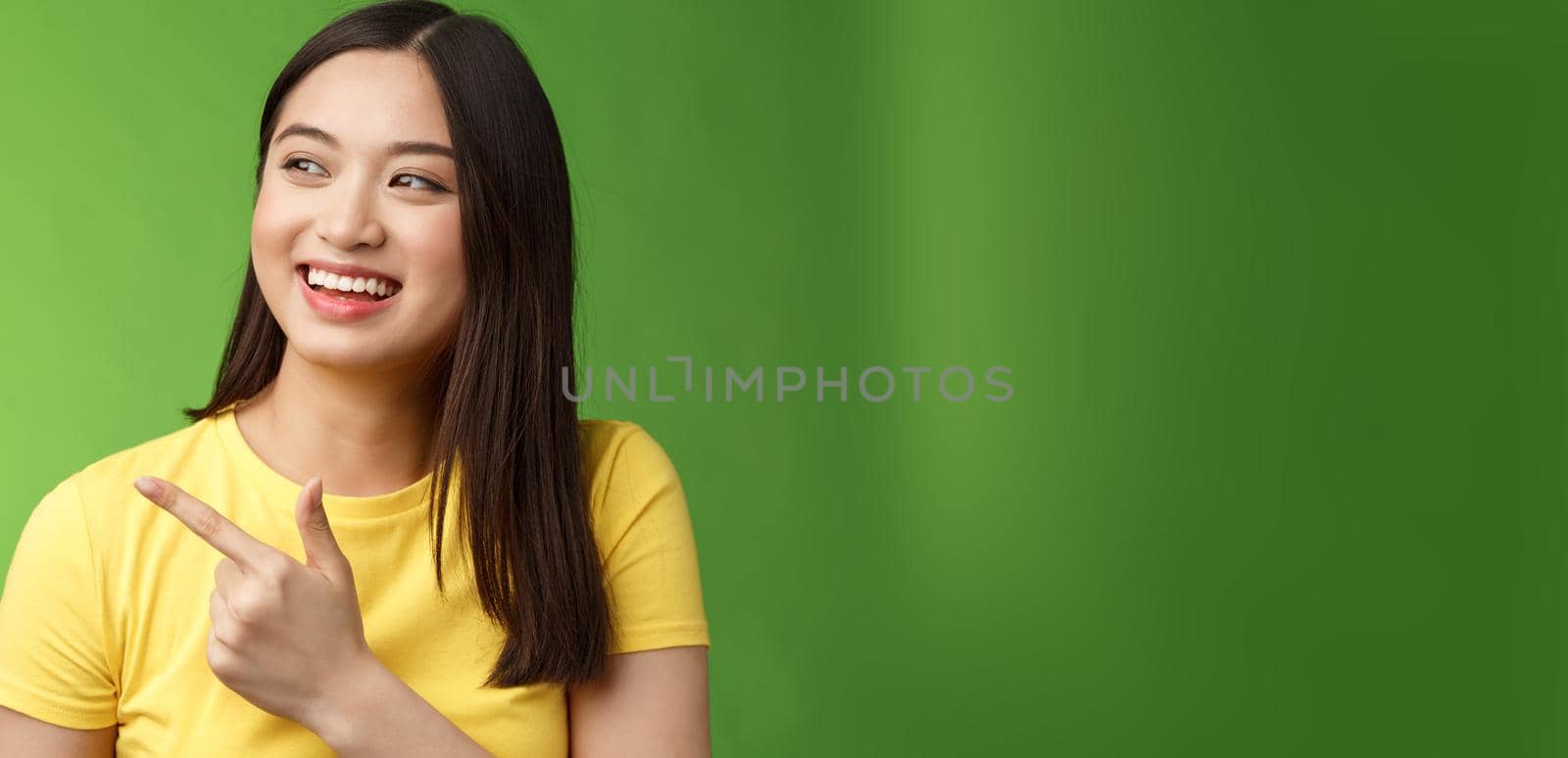 Friendly ougoing cute asian girl having funny conversation, joyfully turn pointing left, indicate awesome cool object, discuss interesting event, chatting carefree, stand green background.