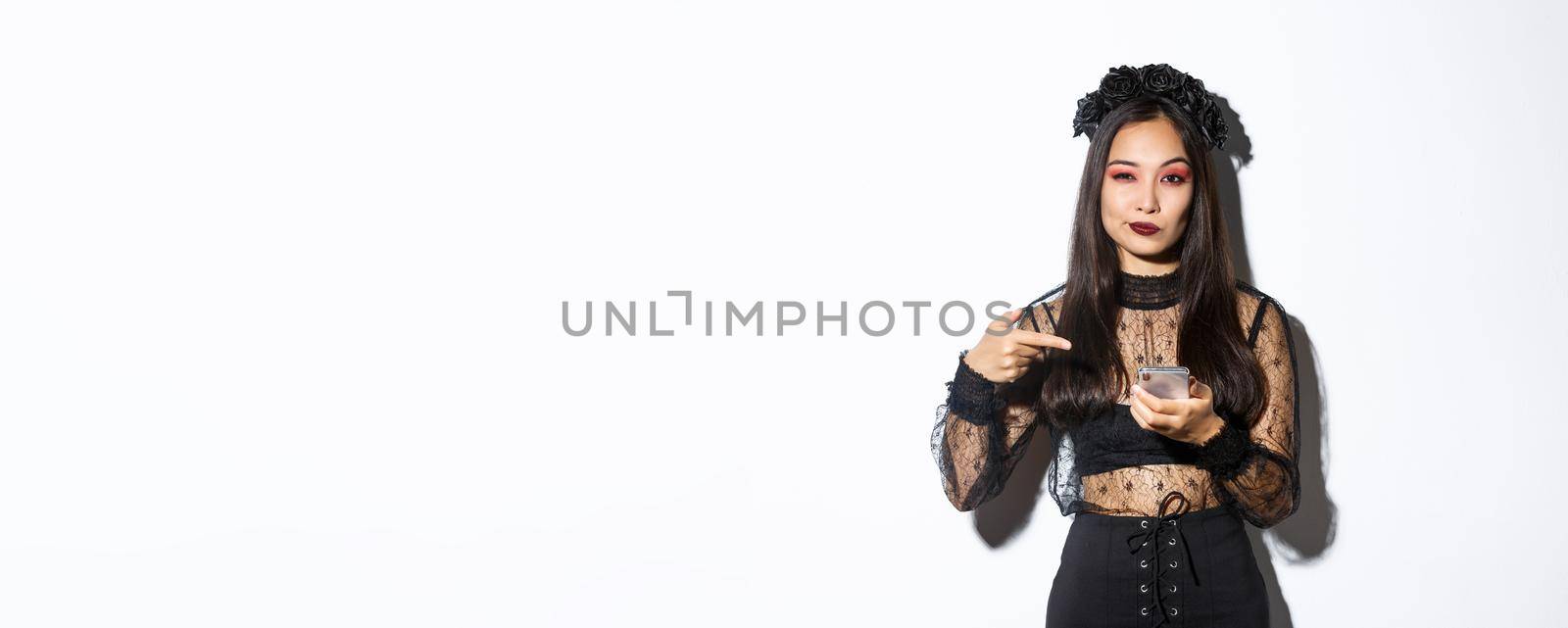 Sassy beautiful asian woman in gothic dress and black wreath pointing finger at mobile phone, showing something about halloween.