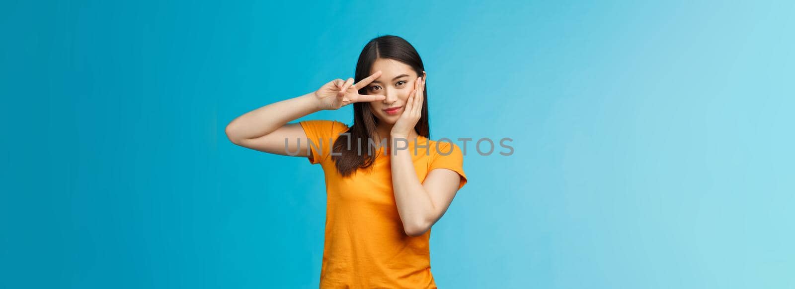 Sassy stylish good-looking asian assertive korean girl show peace victory sign on eye, touch cheek look empowered and motivated, standing cheeky coquettish blue background, wear yellow t-shirt.