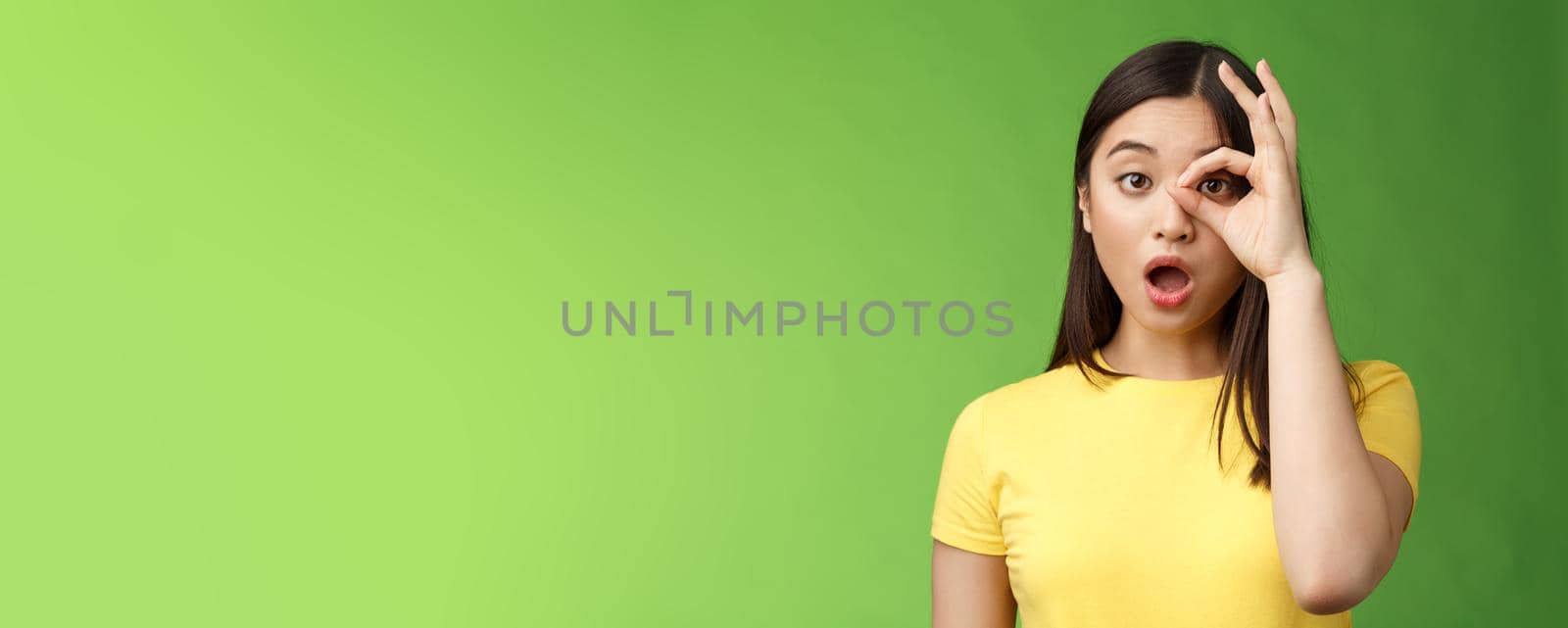 Amazed speechless cute asian young girl drop jaw astonished, look through ok circle sign, drop jaw speechless, impressed look camera, check out incredible promo, stand green background.