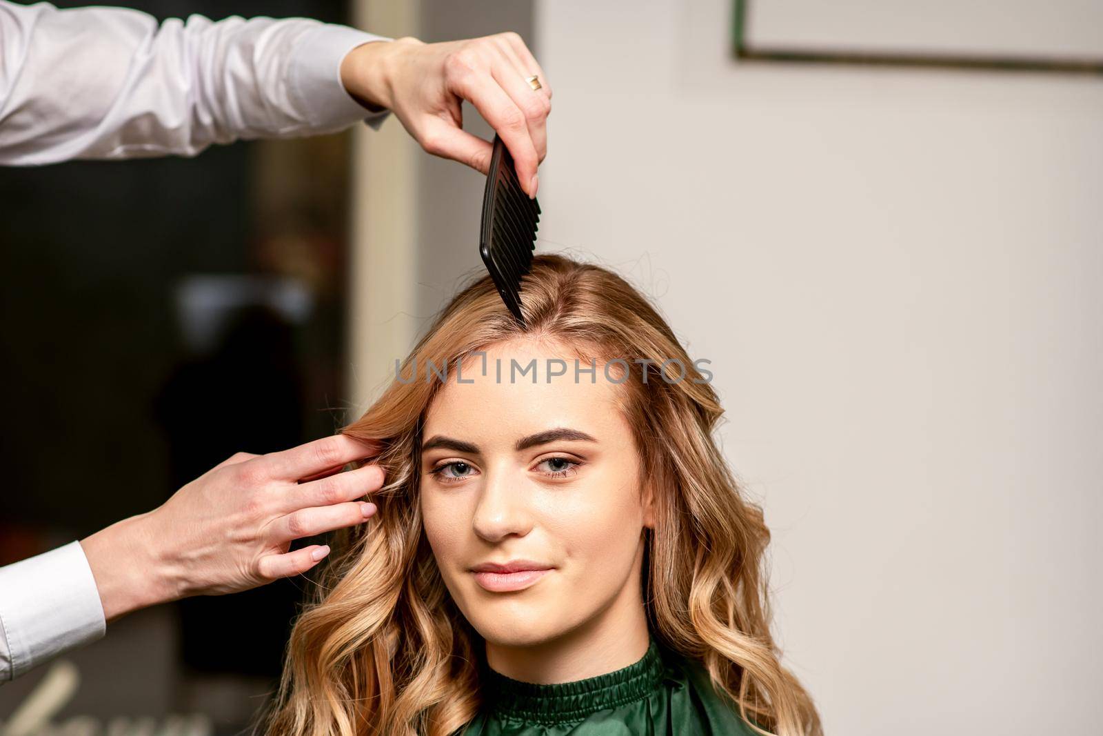 Hairdresser combing long hair of young caucasian woman looking at the camera and smiling in beauty salon. by okskukuruza