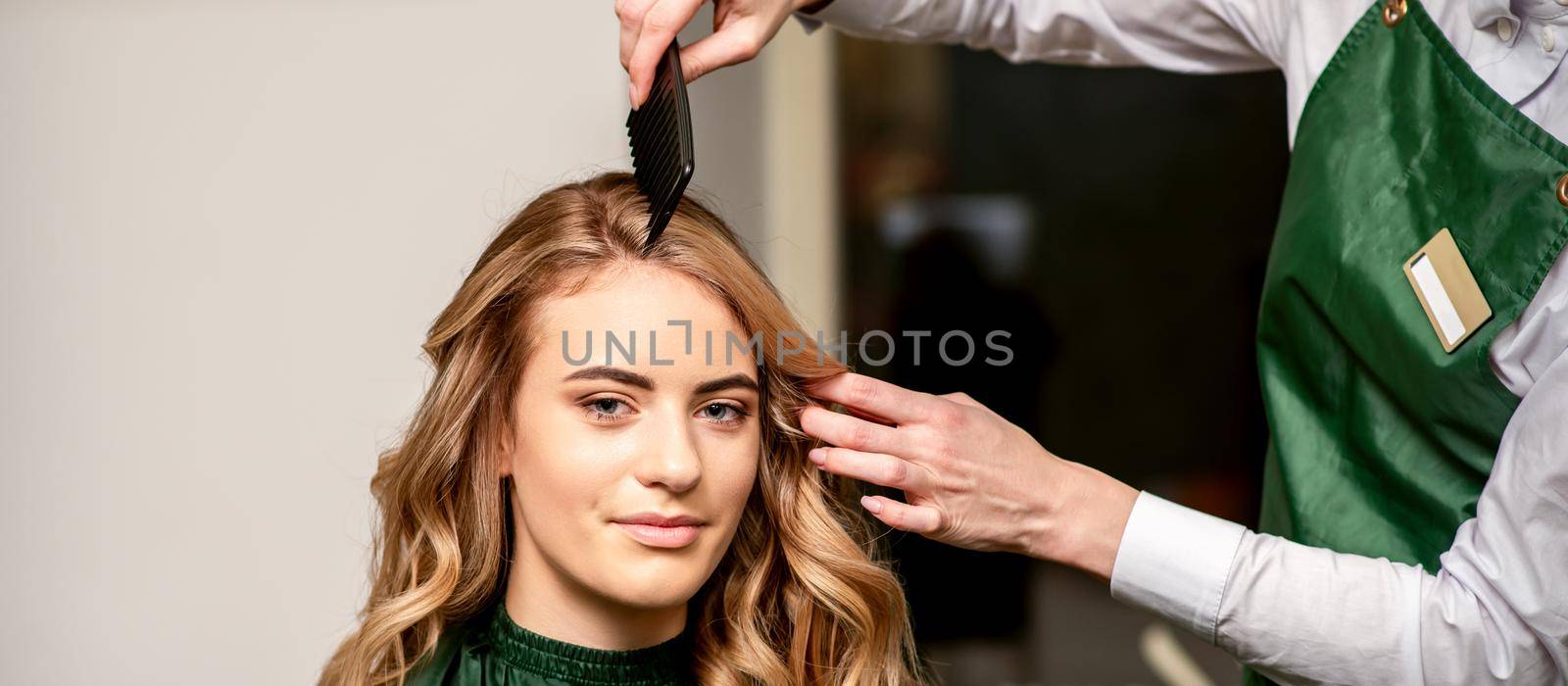 Hairdresser combing long hair of young caucasian woman looking at the camera and smiling in beauty salon. by okskukuruza