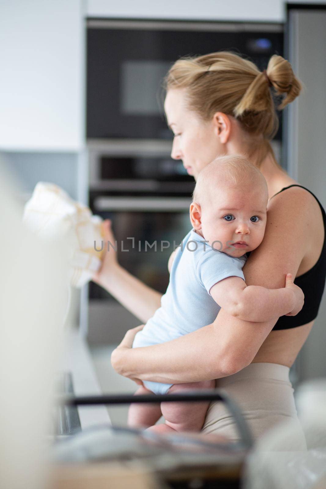 Woman wiping kitchen sink with a cloth after finishing washing the dishes while holding four months old baby boy in her hands by kasto