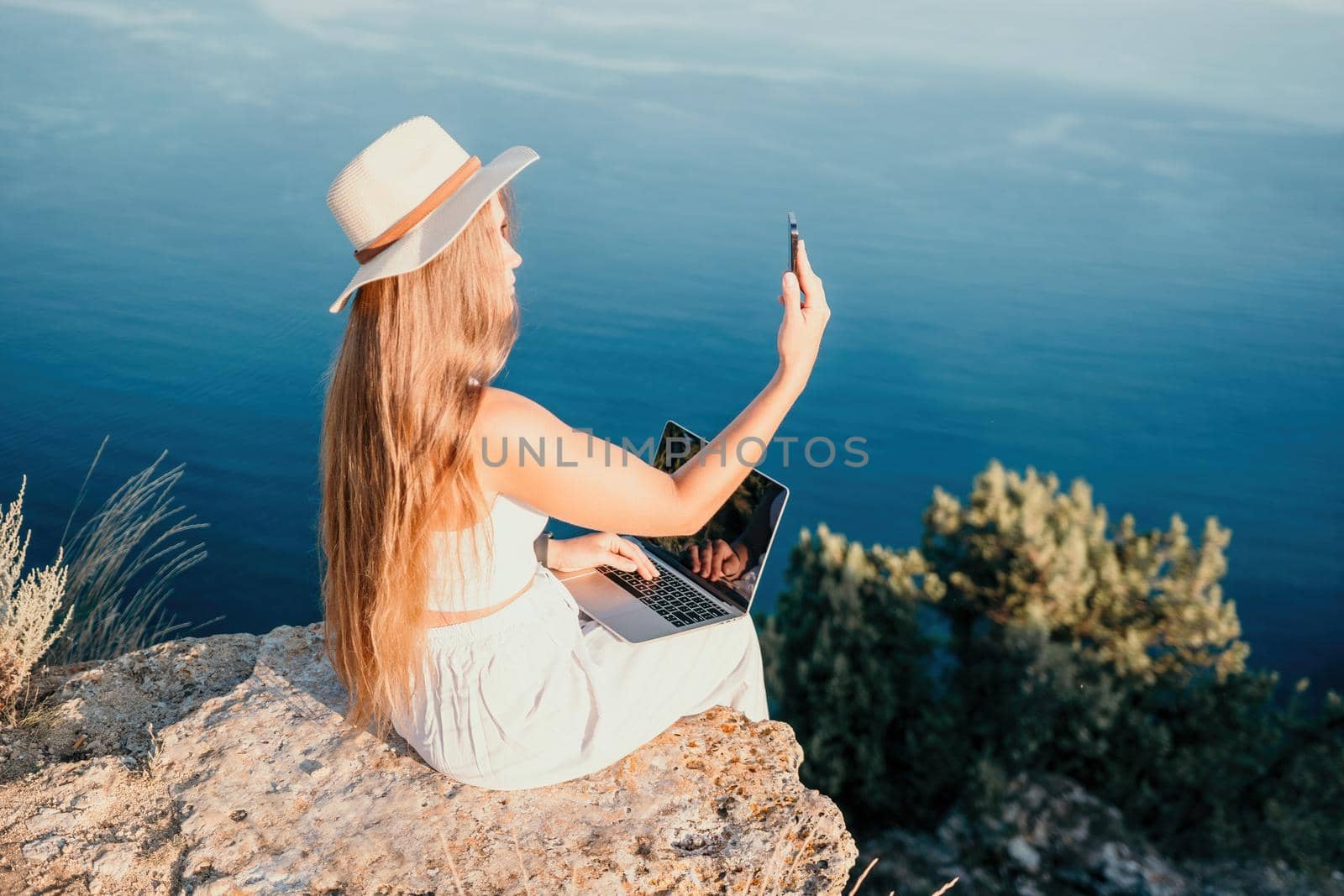 Successful business woman in yellow hat working on laptop by the sea. Pretty lady typing on computer at summer day outdoors. Freelance, travel and holidays concept.