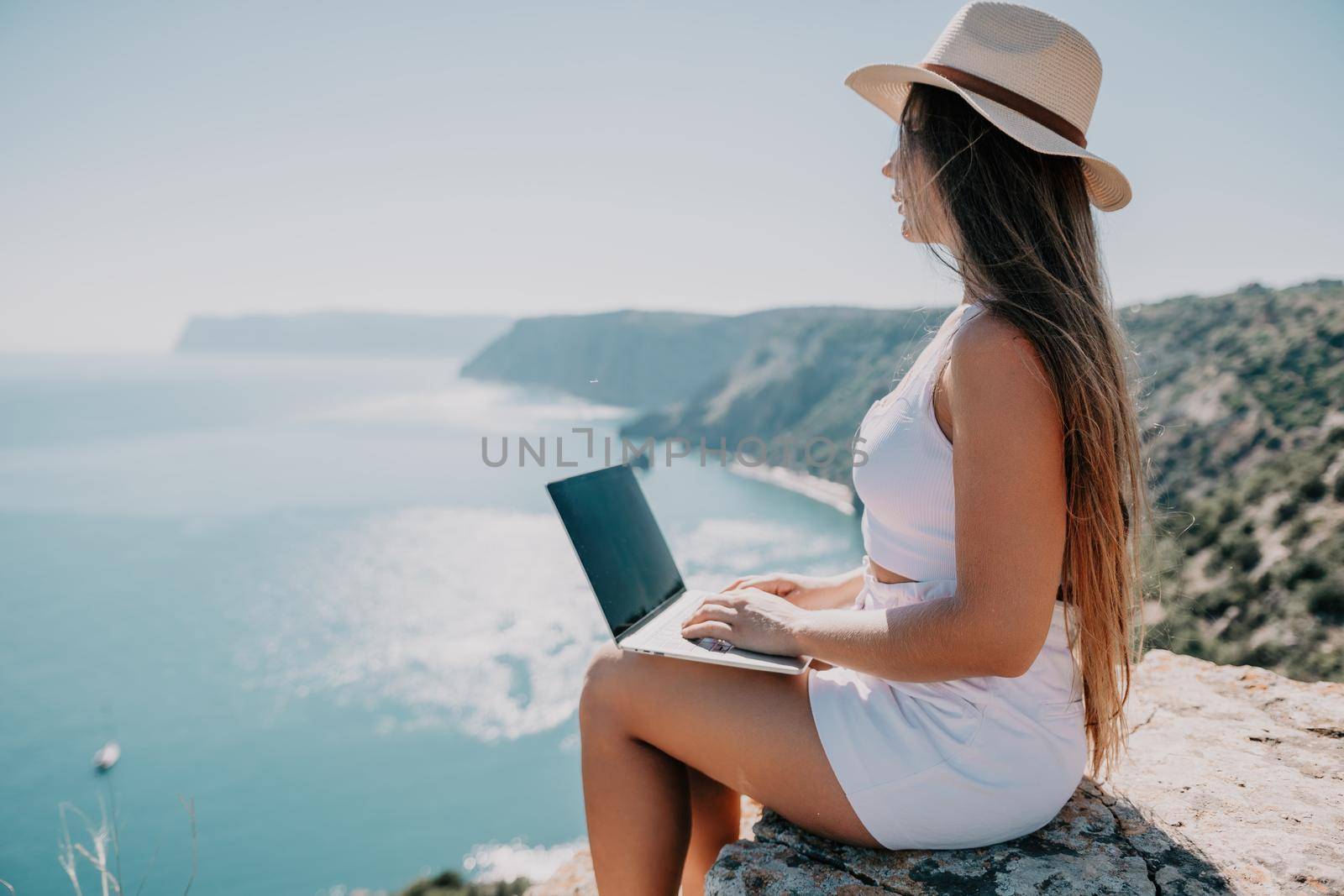 Successful business woman in yellow hat working on laptop by the sea. Pretty lady typing on computer at summer day outdoors. Freelance, travel and holidays concept.