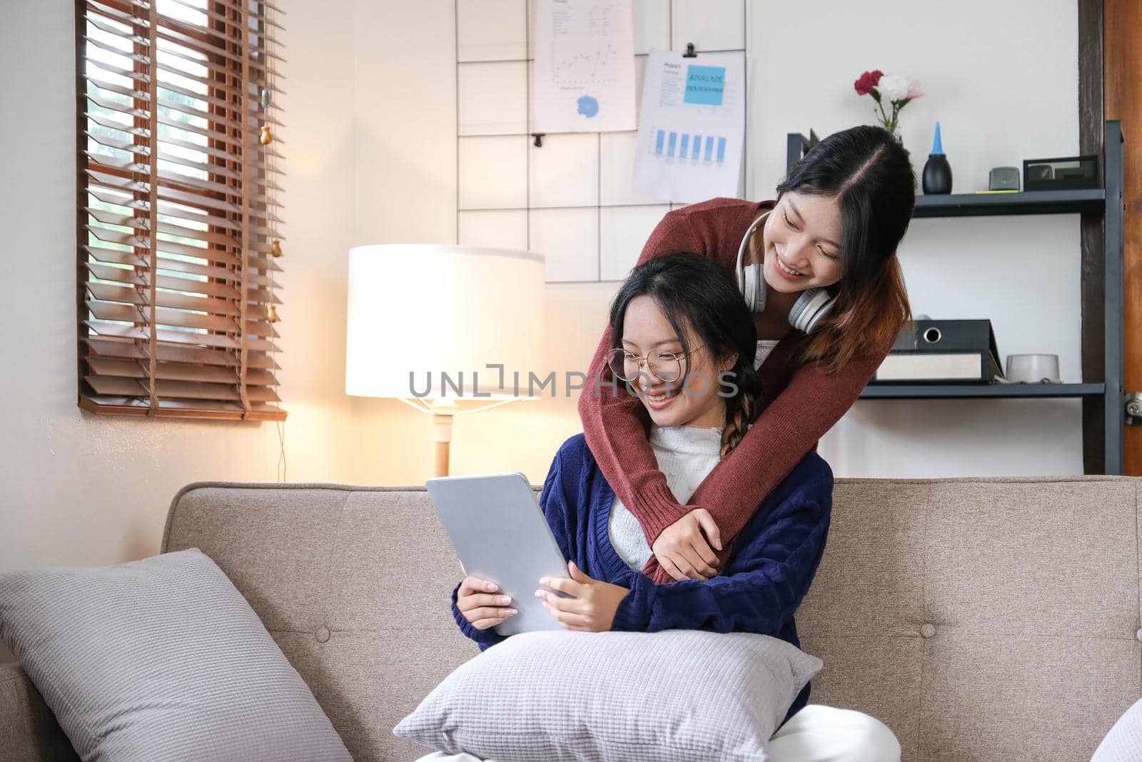 Beautiful Asian lesbian couple hugging their boyfriend in the living room. Two attractive female friends sit on the sofa watching movies on a tablet in the living room. feel happy