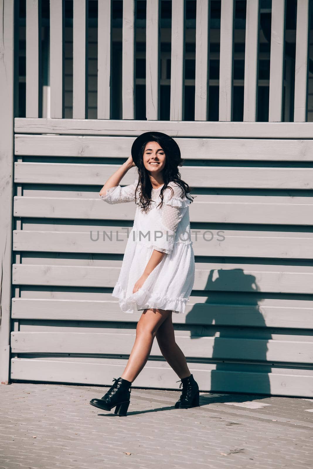 Sunny lifestyle fashion portrait of young stylish hipster woman walking on the street, wearing trendy white dress, black hat and boots. Gray wooden backgrond.