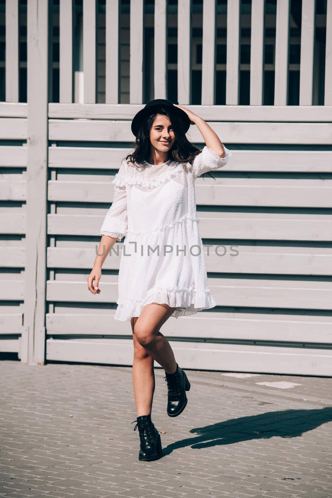 Sunny lifestyle fashion portrait of young stylish hipster woman walking on the street, wearing trendy white dress, black hat and boots. Gray wooden backgrond.