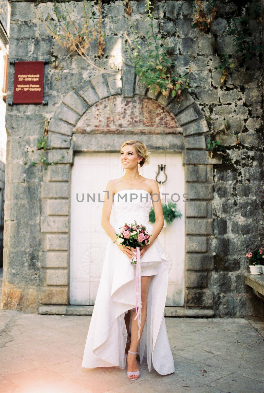 Bride in a white dress with a cutout stands near an ancient stone building. High quality photo