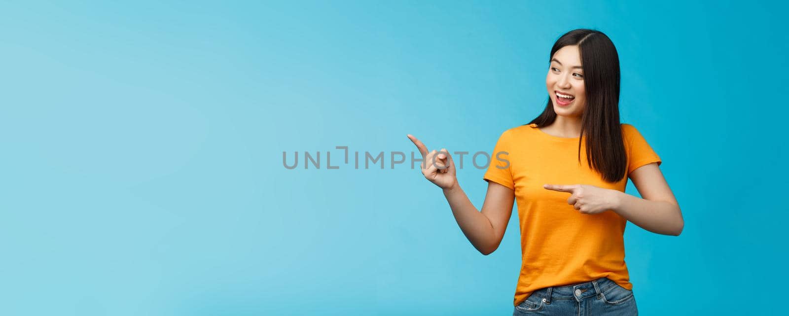 Friendly enthusiastic asian girl curiously looking sideways, pointing left interesting object, grinning joyfully discuss new opened store, stand blue background carefree lively talking by Benzoix
