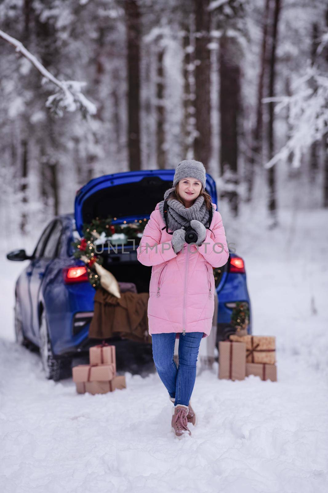 A woman in a winter snow-covered forest in the trunk of a car decorated with Christmas decor. A female photographer holds a camera in her hands.