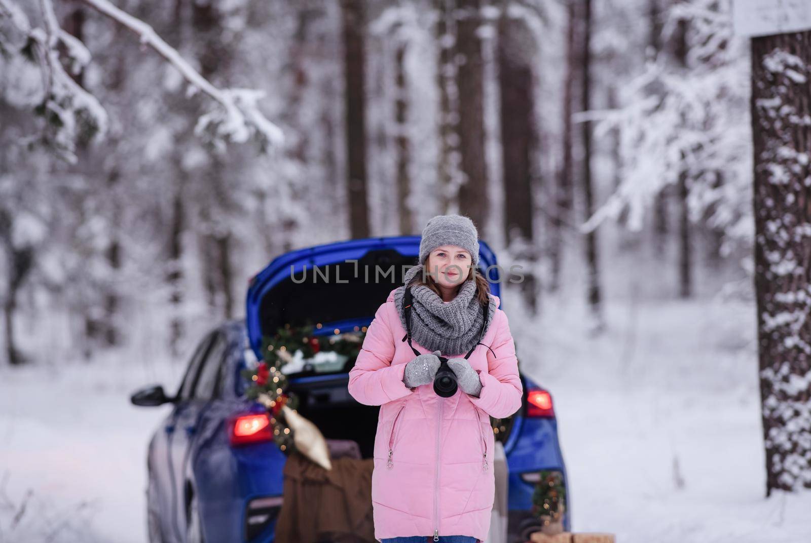 A woman in a winter snow-covered forest in the trunk of a car decorated with Christmas decor. A female photographer holds a camera in her hands.