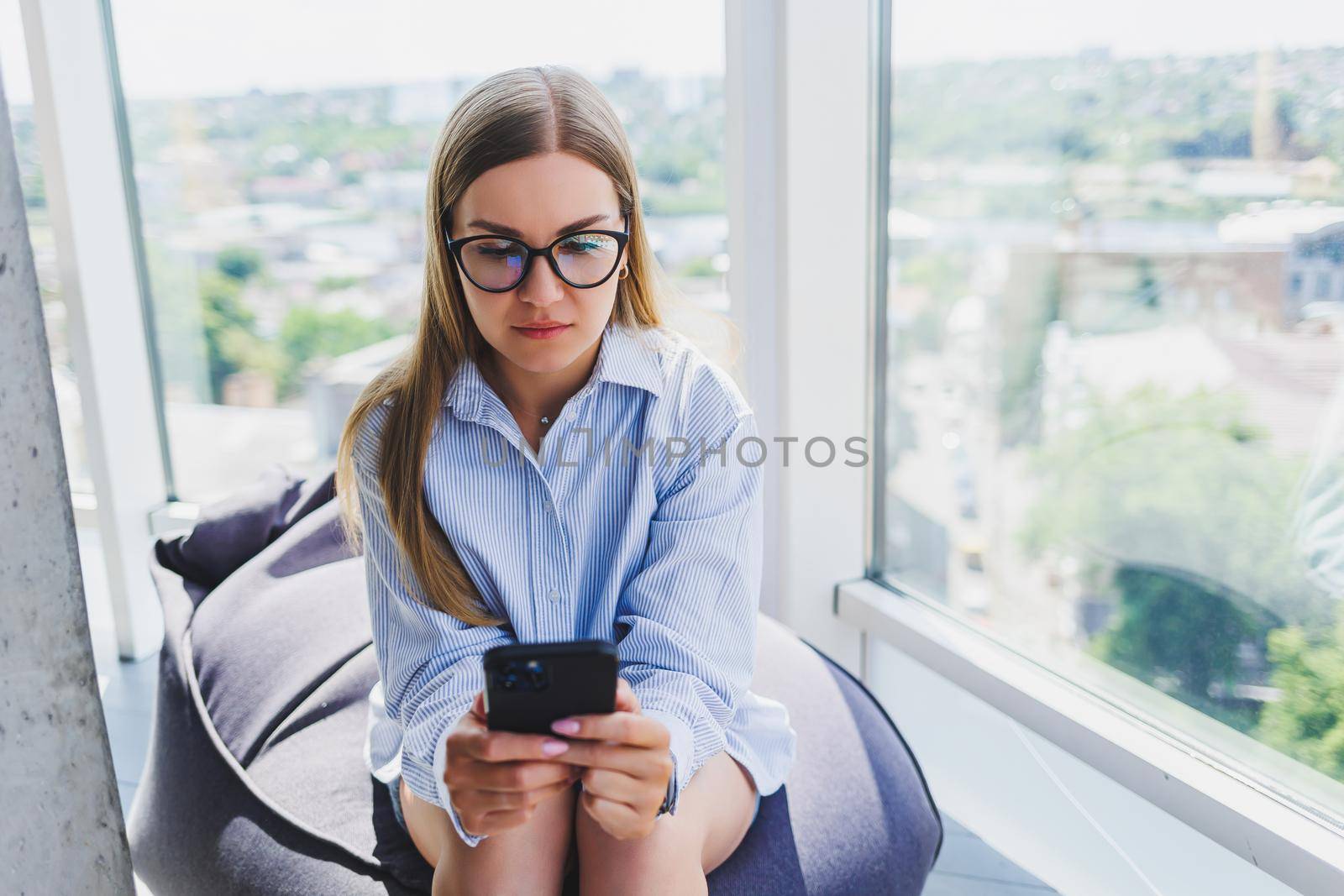 Happy freelancer girl in classic glasses looking at phone and smiling while sitting in modern coworking space, carefree millennial woman in glasses enjoying leisure time for communication