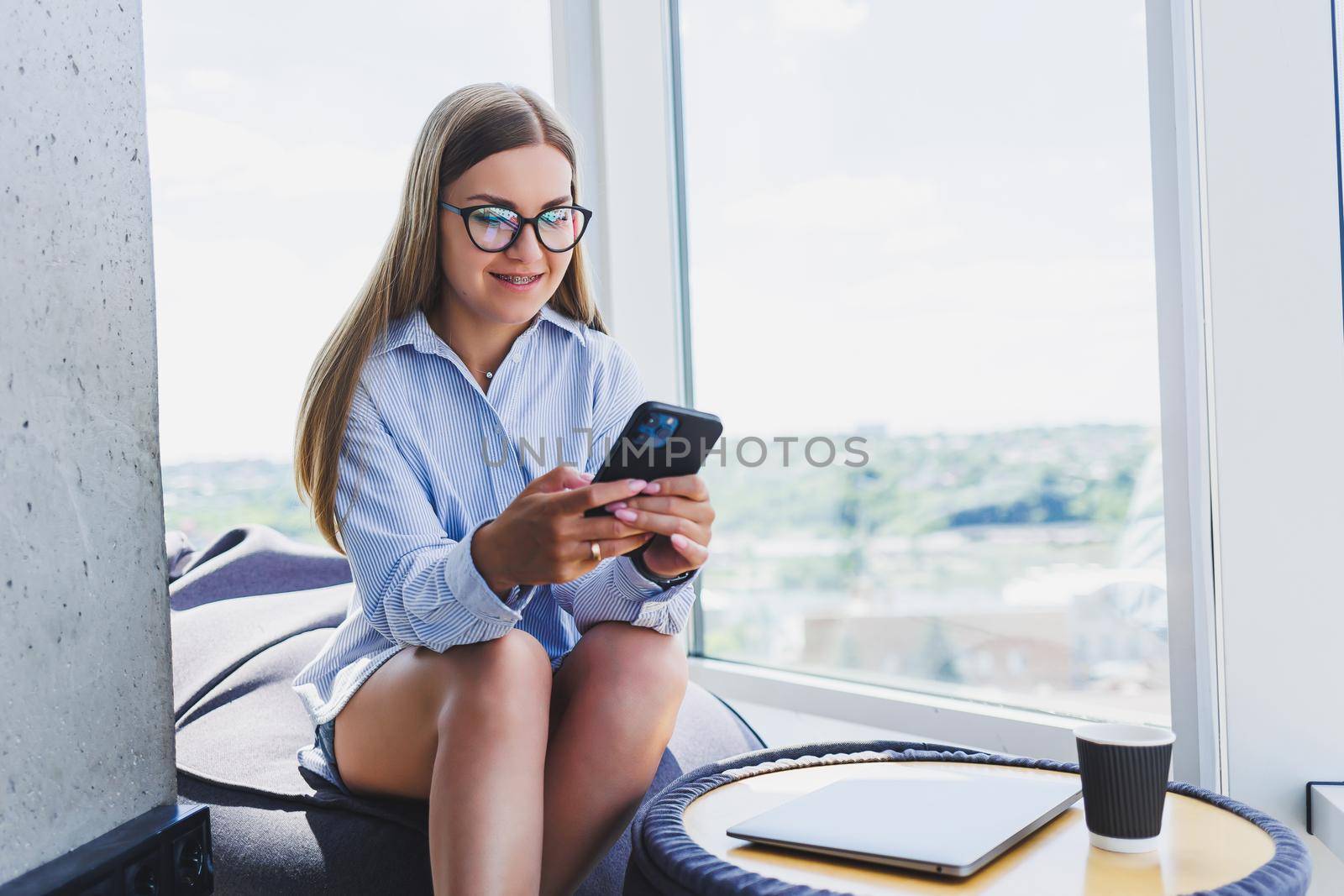 Happy freelancer girl in classic glasses looking at phone and smiling while sitting in modern coworking space, carefree millennial woman in glasses enjoying leisure time for communication by Dmitrytph