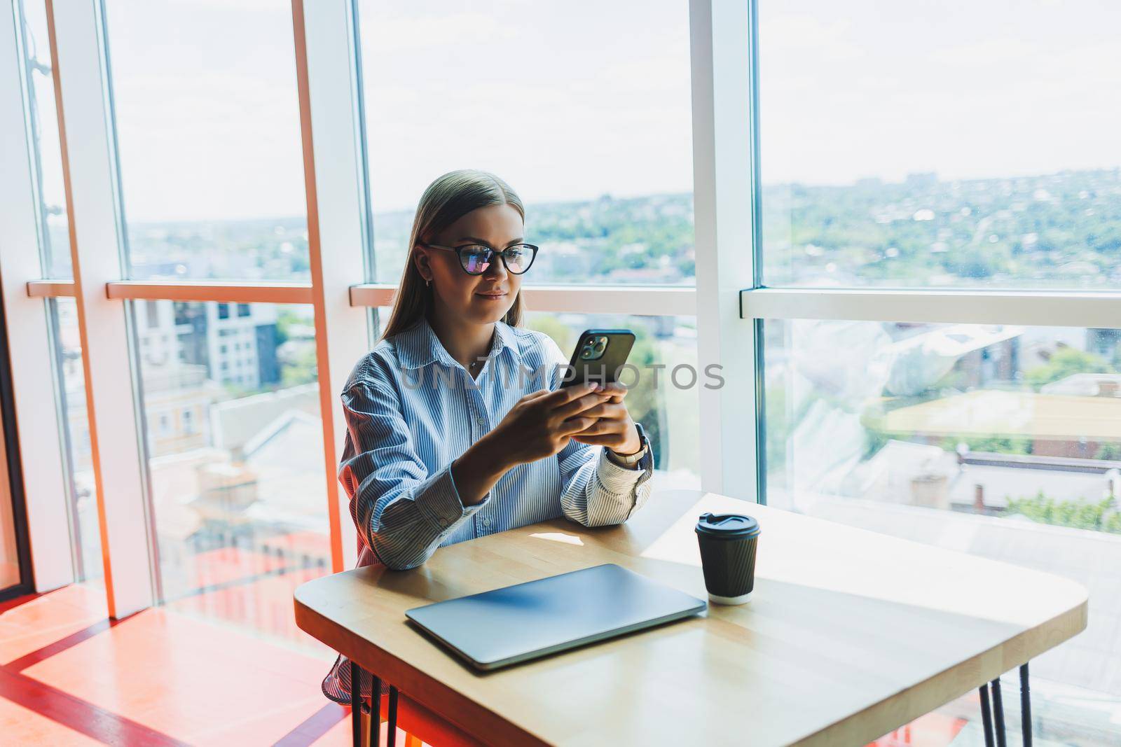 Happy freelancer girl in classic glasses looking at phone and smiling while sitting in modern coworking space, carefree millennial woman in glasses enjoying leisure time for communication by Dmitrytph