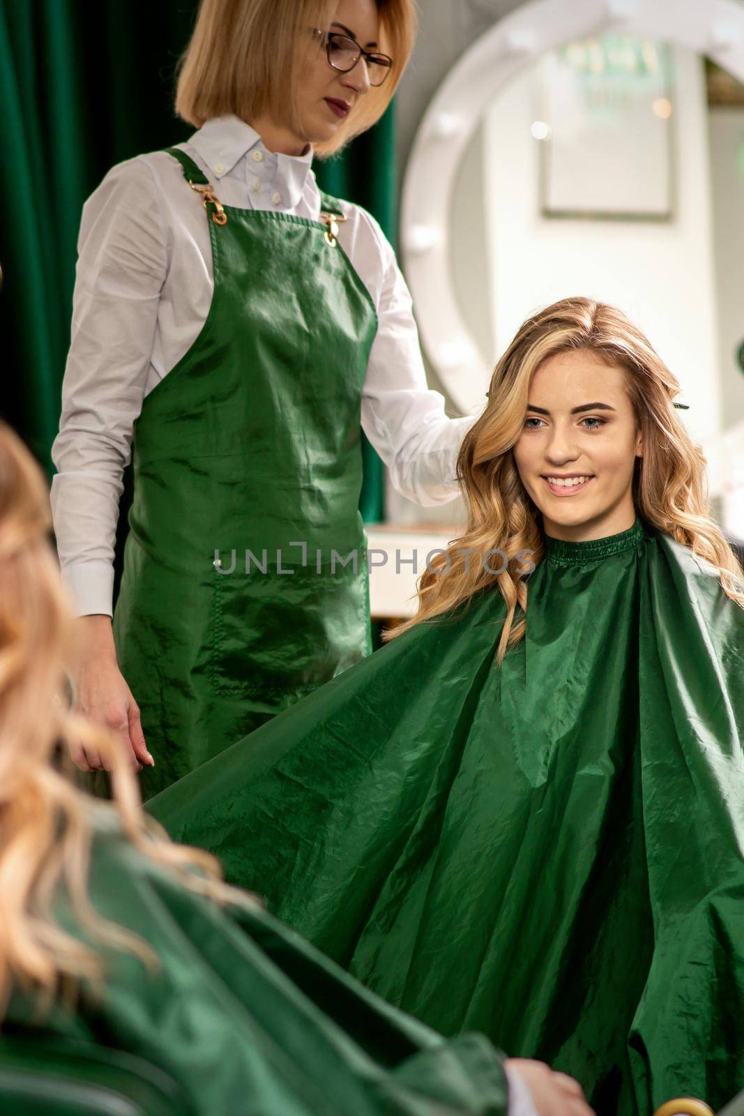 Hairdresser combing long hair of young caucasian woman looking and smiling in the mirror in beauty salon