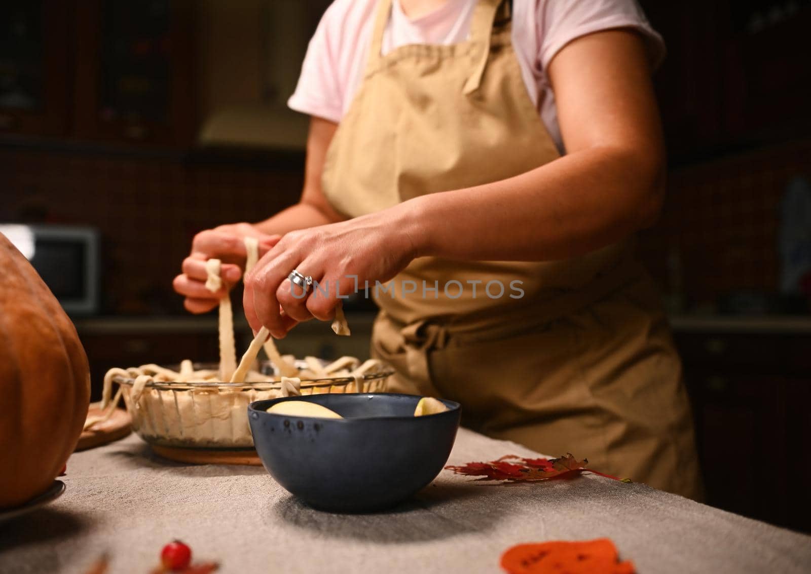 Cropped view of pastry chef in beige apron standing at table with linen tablecloth, decorating homemade pie for Thanksgiving dinner, holding pastry strips, making lattice pattern on top of pumpkin pie