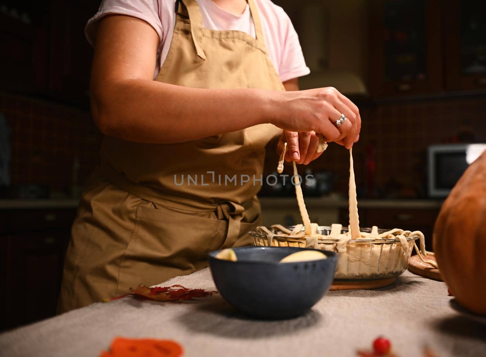 Close-up of a housewife in beige chef's apron, decorating the classic American homemade festive pie with a crunchy pastry lattice. Thanksgiving Day. Making tasty pumpkin apple pie with flaky crust.