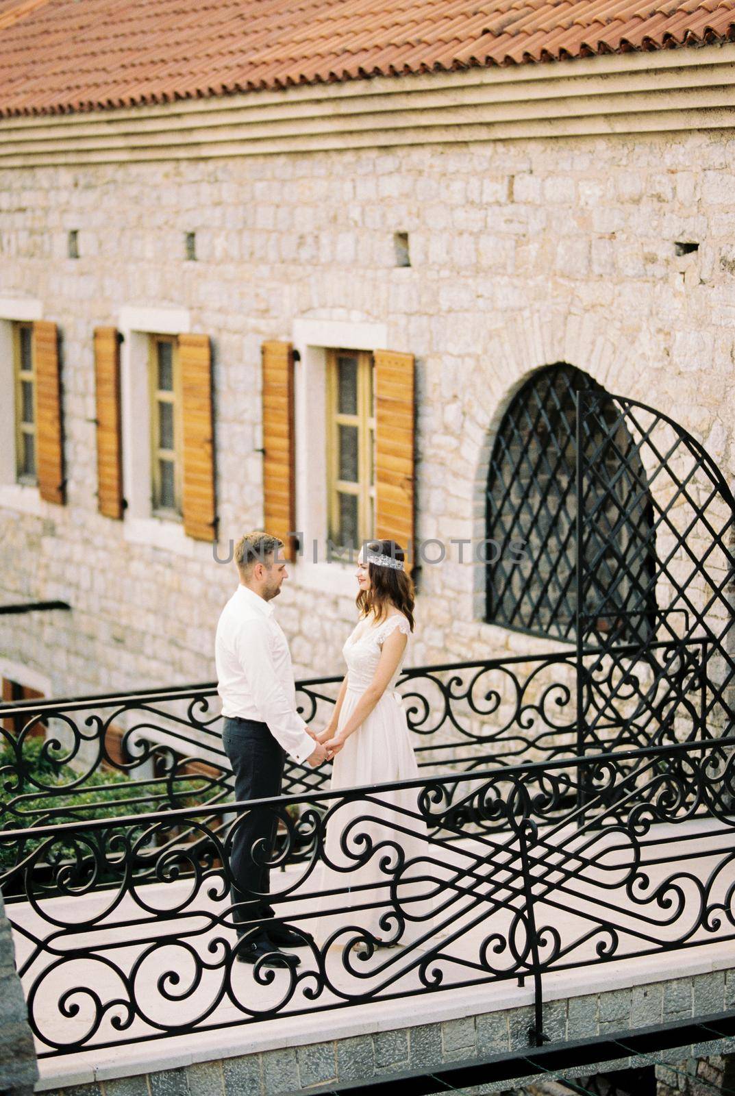 Bride and groom stand holding hands on the bridge near the old stone building. High quality photo