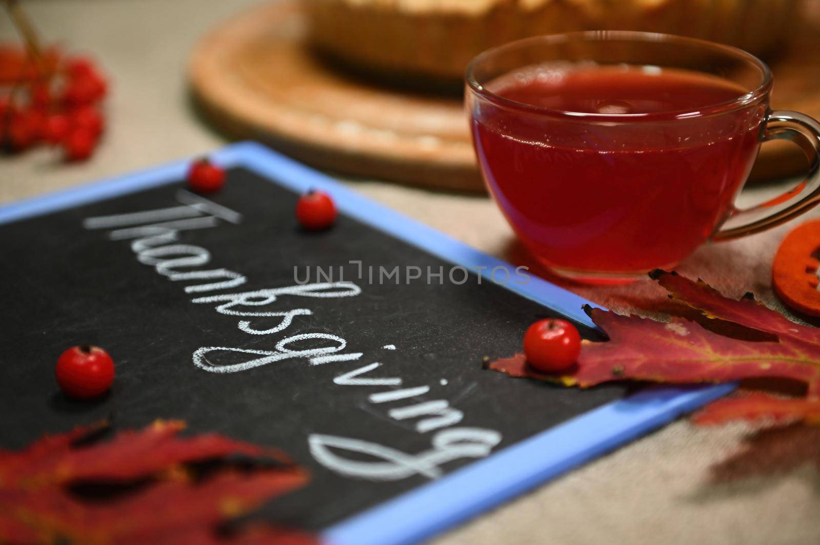 Selective focus. Still life with a glass teacup with healthy antioxidant hot herbal drink, blurred foreground with dry fallen autumn maple leaves and a blackboard with lettering Thanksgiving Day.