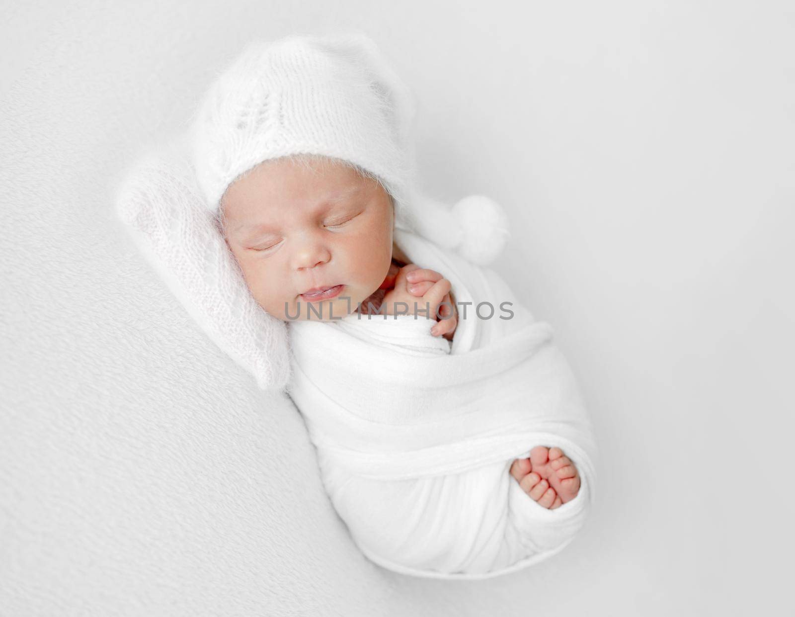 Newborn baby girl swaddled in white fabric and wearing hat sleeping. Infant child kid studio portrait