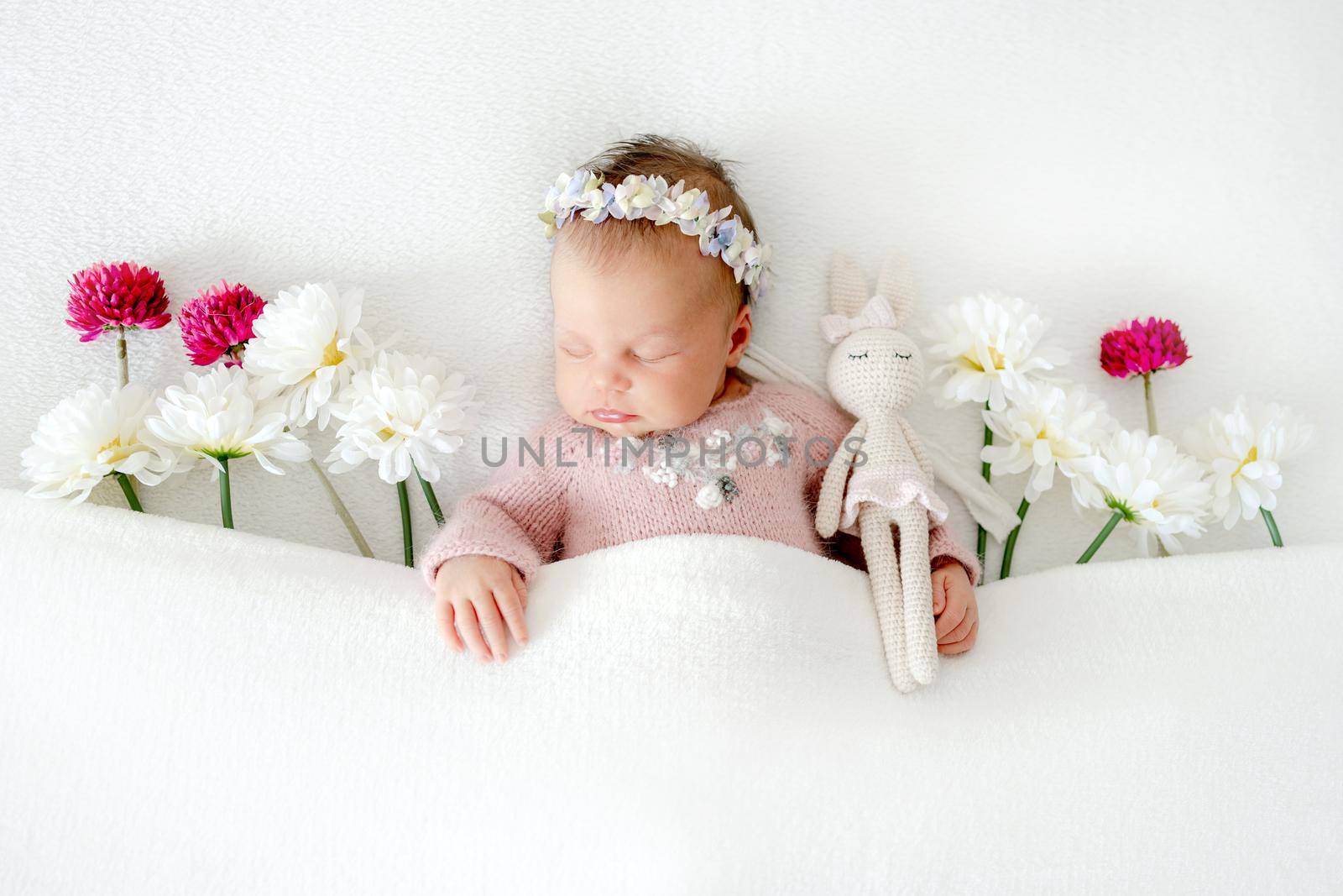 Newborn baby girl wearing wreath sleeping in bed decorated with white and pink flowers. Infant child kid spring blossom studio portrait