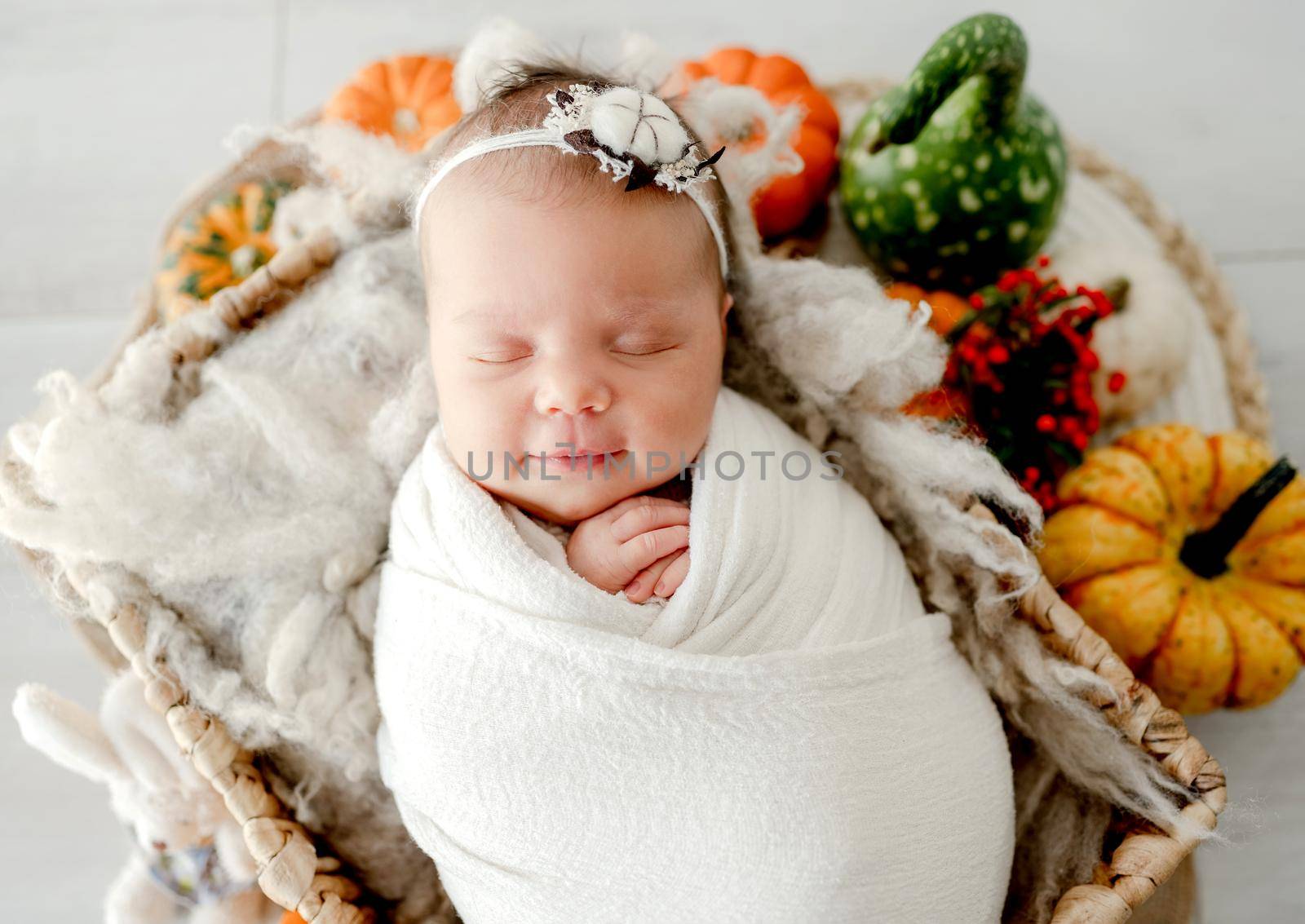 Newborn baby girl swaddled in white fabric sleeping in basket decorated with pumpkins. Infant child kid napping on fur autumn studio portrait