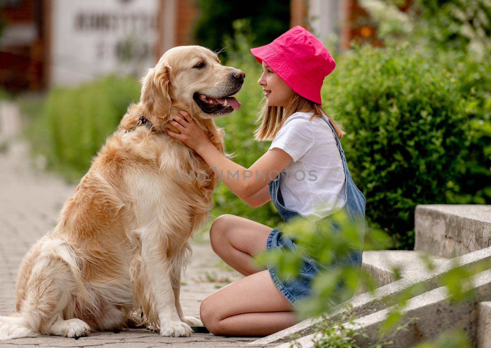 Preteen girl wearing hat hugging golden retriever dog sitting outdoors in summertime. Pretty kid petting fluffy doggy pet in city