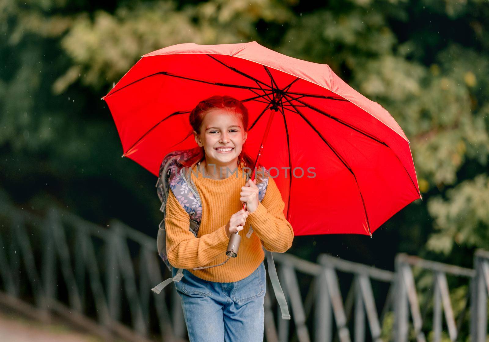 School girl with backpack and umbrella outdoors. Pretty child kid walking in rainy weather