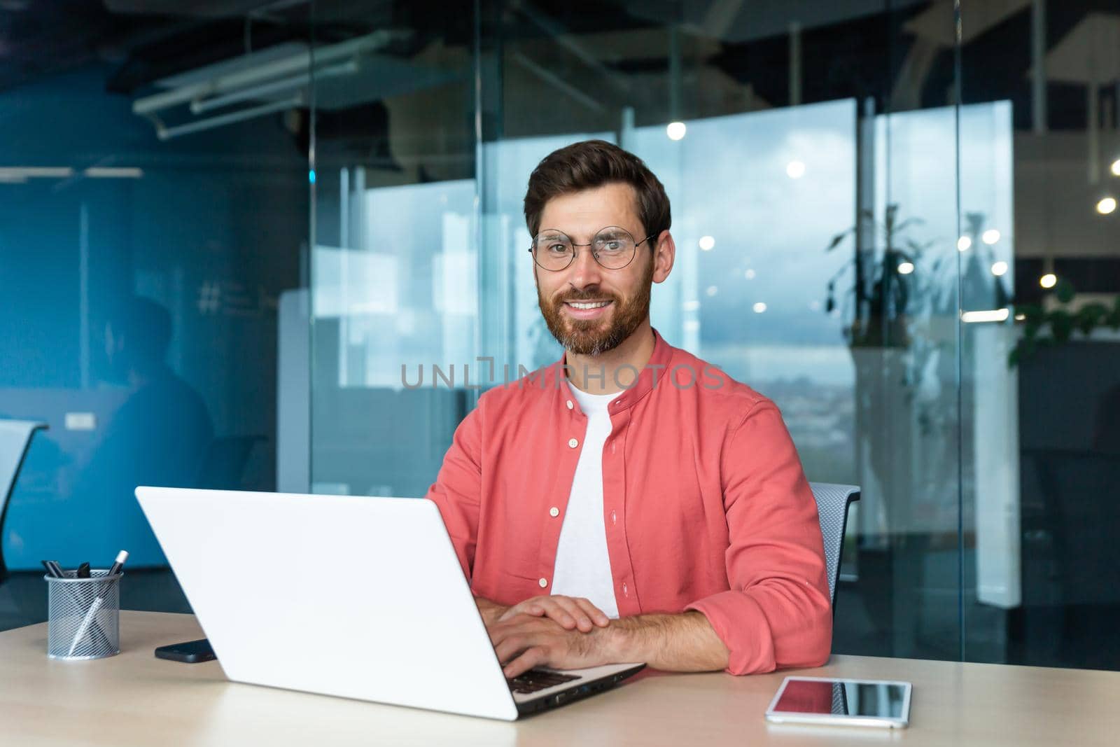 Portrait of mature businessman freelancer startup, bearded man smiling and looking at camera, business owner working inside modern office building wearing red shirt and glasses.