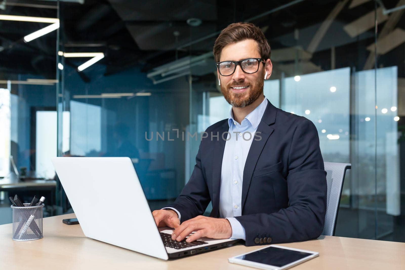 Portrait of mature boss inside office with laptop, successful and satisfied investor manager looking at camera and smiling man in glasses and business suit, investor with beard sitting on chair